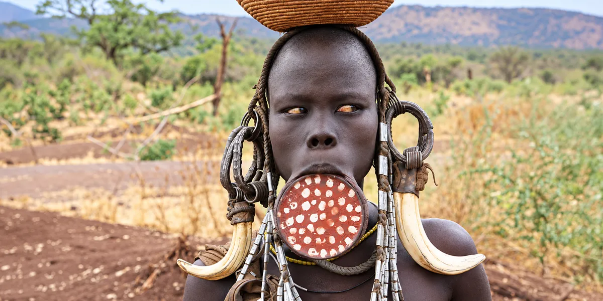 A Mursi woman in southern Ethiopia wearing a large lip plate and curved horn ornaments, with a woven basket on her head.