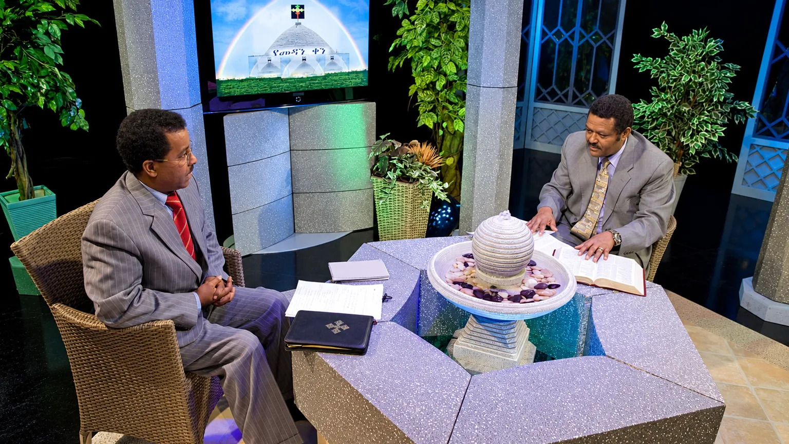 Two men discuss Bible teachings in a Christian television studio, with an open Bible on the table.