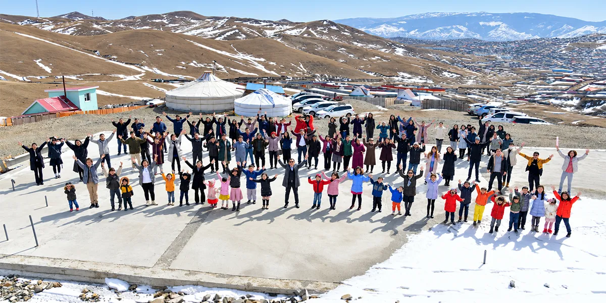 Adults and children wave together outdoors in Mongolia, with traditional gers and snow-covered mountains in the background.